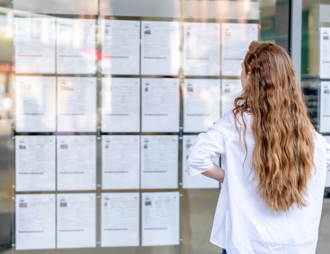A person with long hair stands in front of a glass window, carefully reviewing job listings that are displayed on the wall outside a public space.