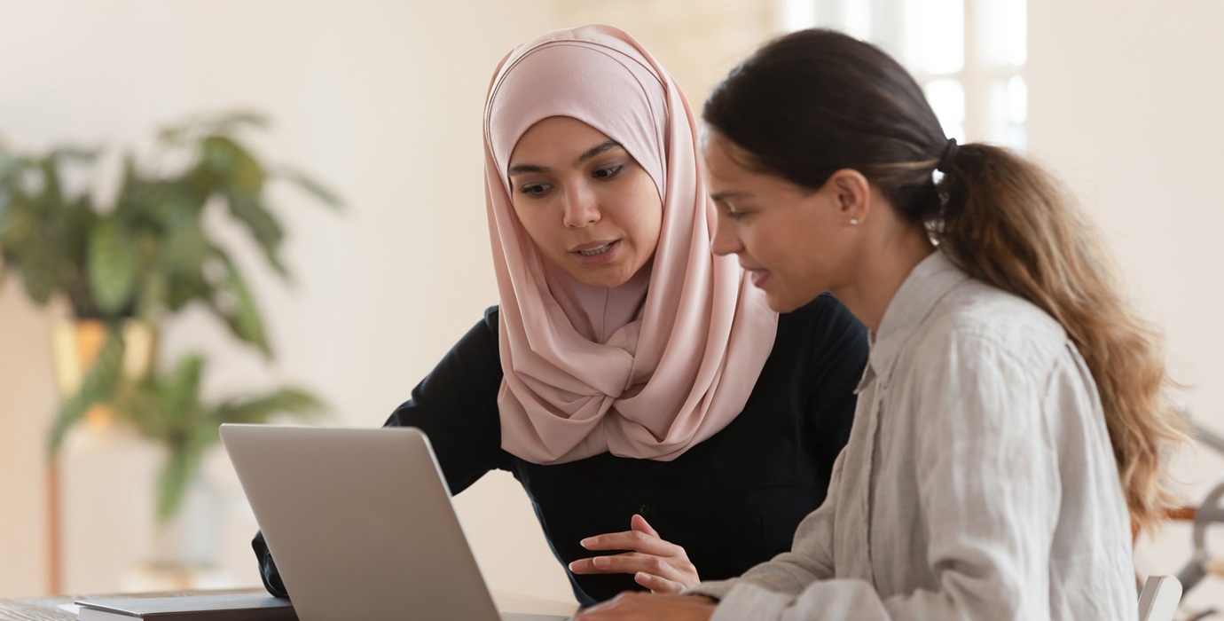 An employee training an intern on a laptop while they sit at an office desk. 