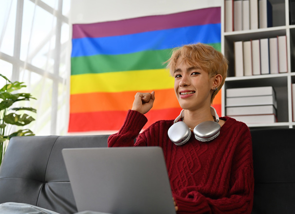 Young person at laptop fist pumping with pride flag behind