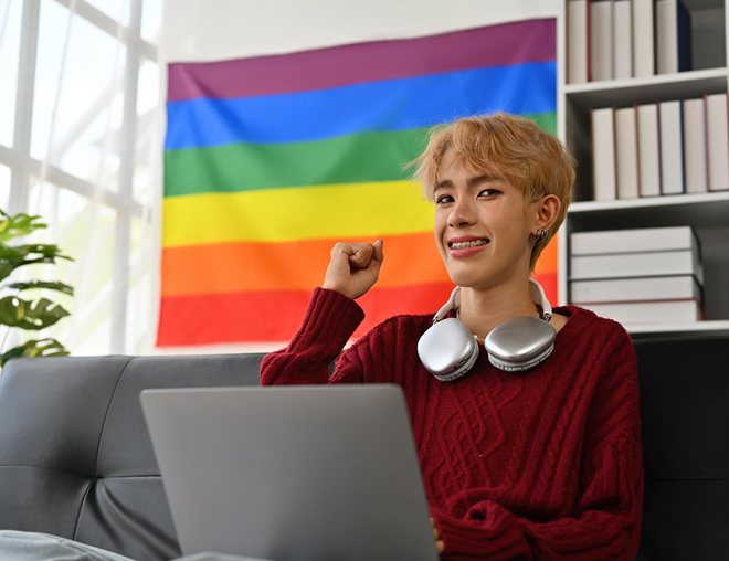 Young person at laptop fist pumping with pride flag behind