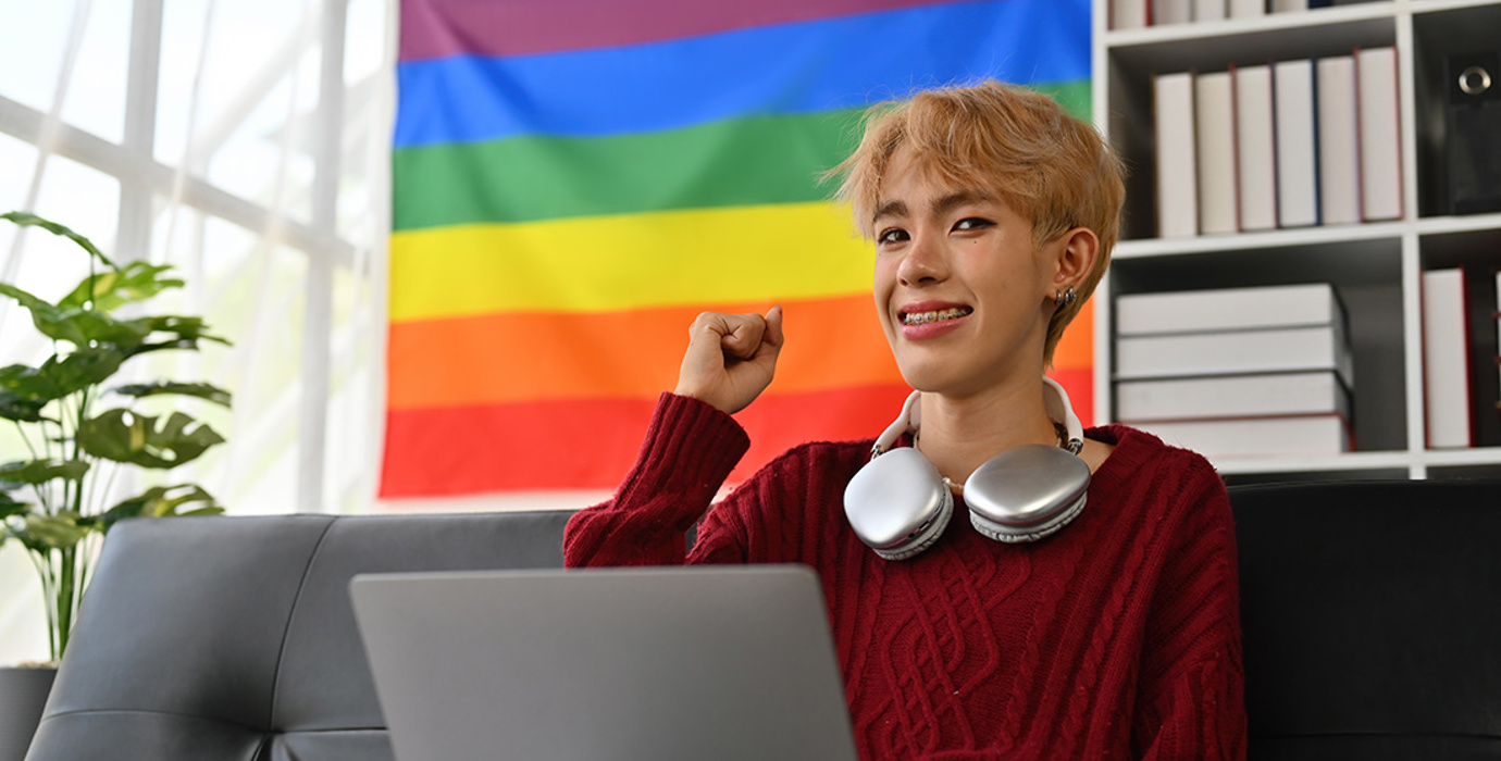 Young person at laptop fist pumping with pride flag behind