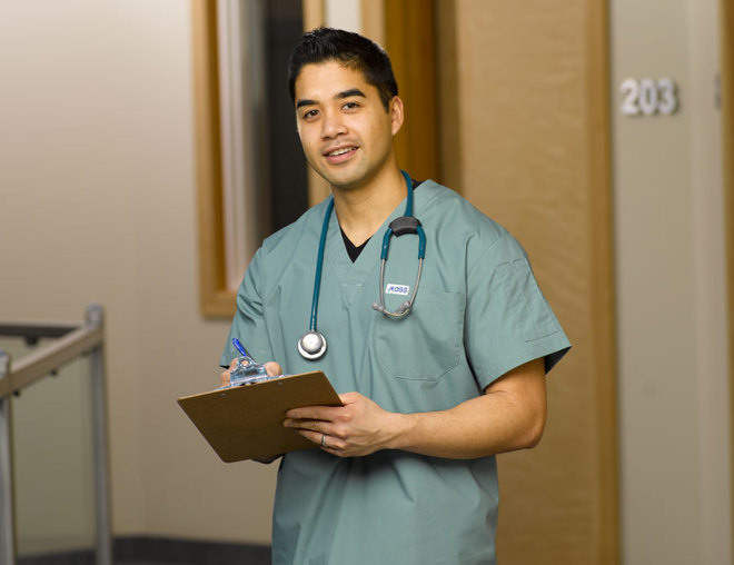 Nurse wearing scrubs with stethoscope around neck, carrying a clipboard stands in a hospital hallway