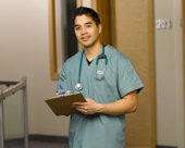 Nurse wearing scrubs with stethoscope around neck, carrying a clipboard stands in a hospital hallway
