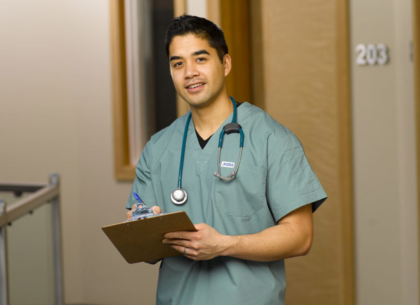Nurse wearing scrubs with stethoscope around neck, carrying a clipboard stands in a hospital hallway
