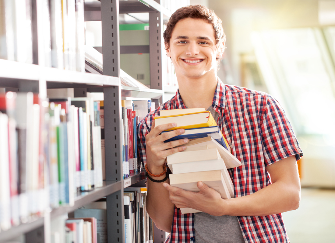 Youth holding a stack of books while leaning against a library shelf