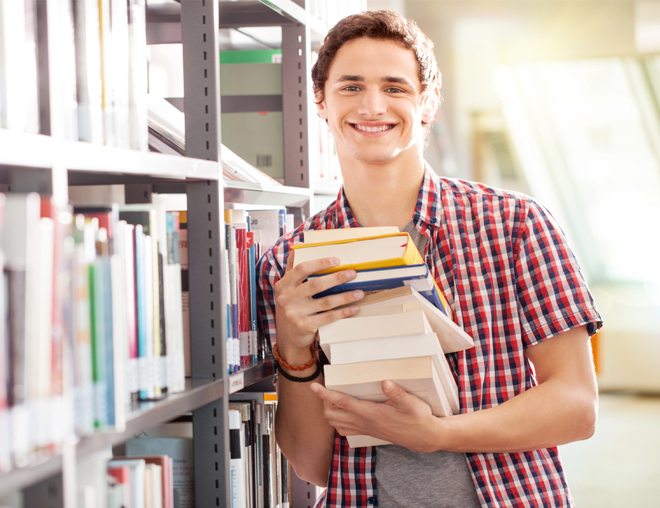Youth holding a stack of books while leaning against a library shelf