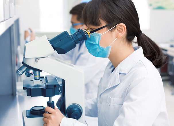 Person in a laboratory wearing a lab coat and mask using a microscope