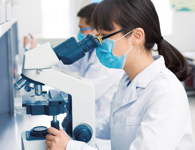 Person in a laboratory wearing a lab coat and mask using a microscope