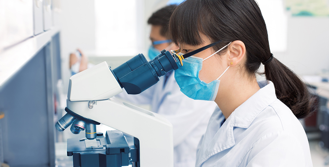 Person in a laboratory wearing a lab coat and mask using a microscope