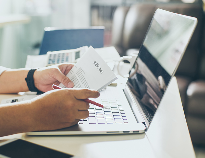 Hands holding a resume and pencil on top of an open laptop