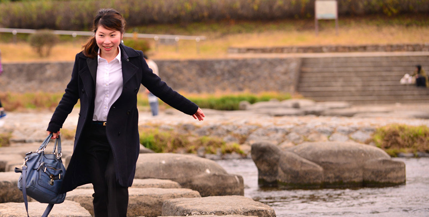 Person wearing office attire carrying a bag stepping on stones across a river