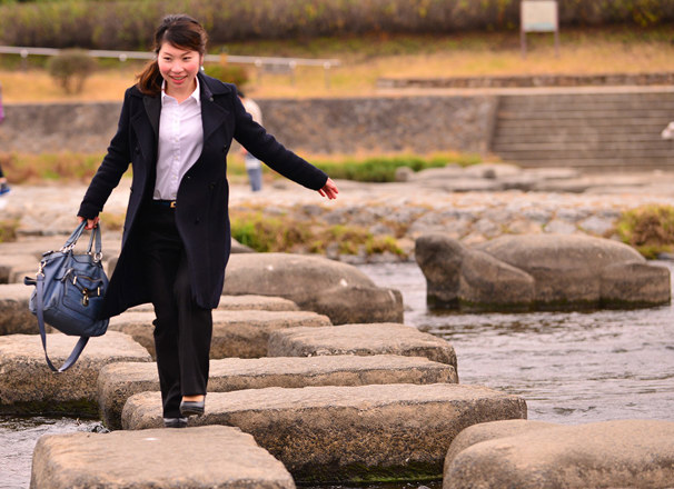 Person wearing office attire carrying a bag stepping on stones across a river