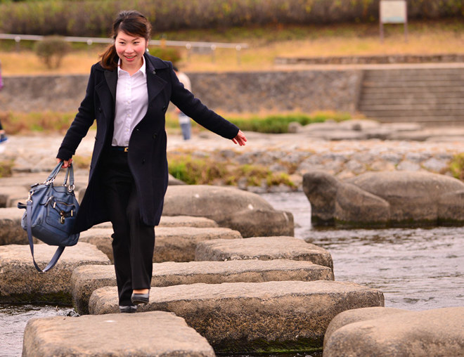 Person wearing office attire carrying a bag stepping on stones across a river