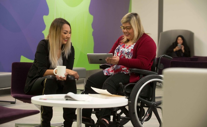 Student holding a tablet while chatting with a friend in a campus common room