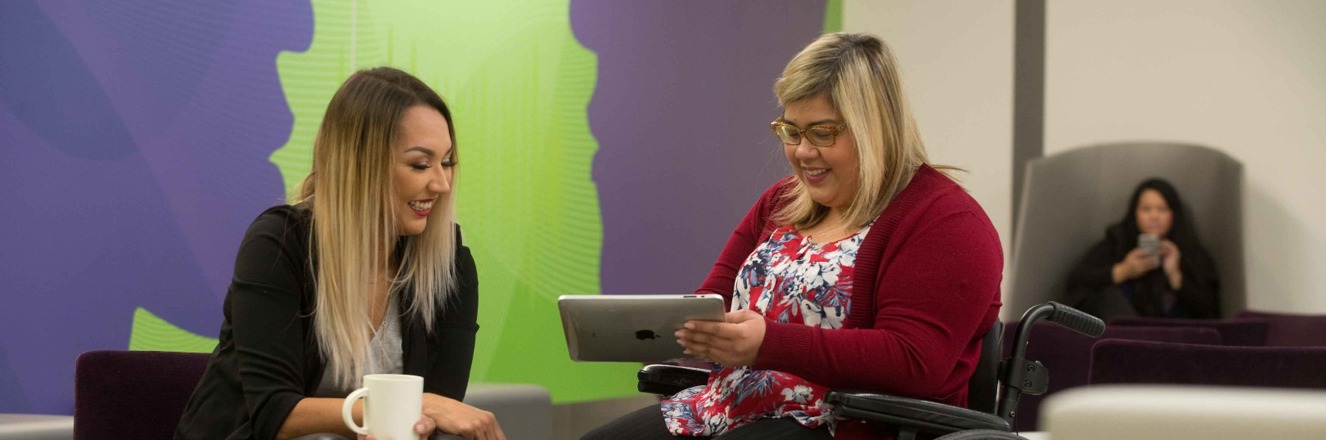 Student holding a tablet while chatting with a friend in a campus common room