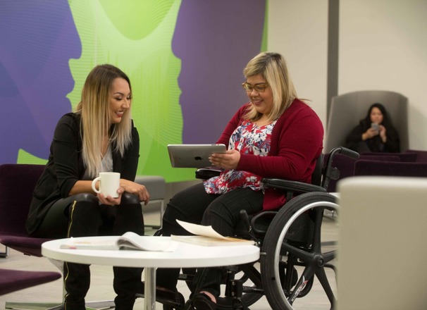Student holding a tablet while chatting with a friend in a campus common room