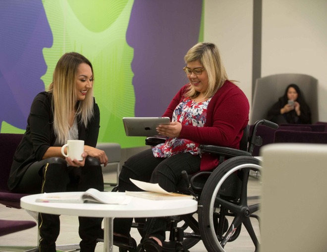 Student holding a tablet while chatting with a friend in a campus common room