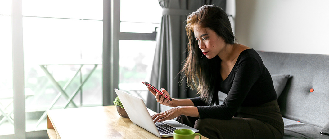 Person sitting on a couch in the living room looking at a laptop and smartphone