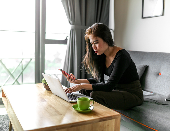 Person sitting on a couch in the living room looking at a laptop and smartphone