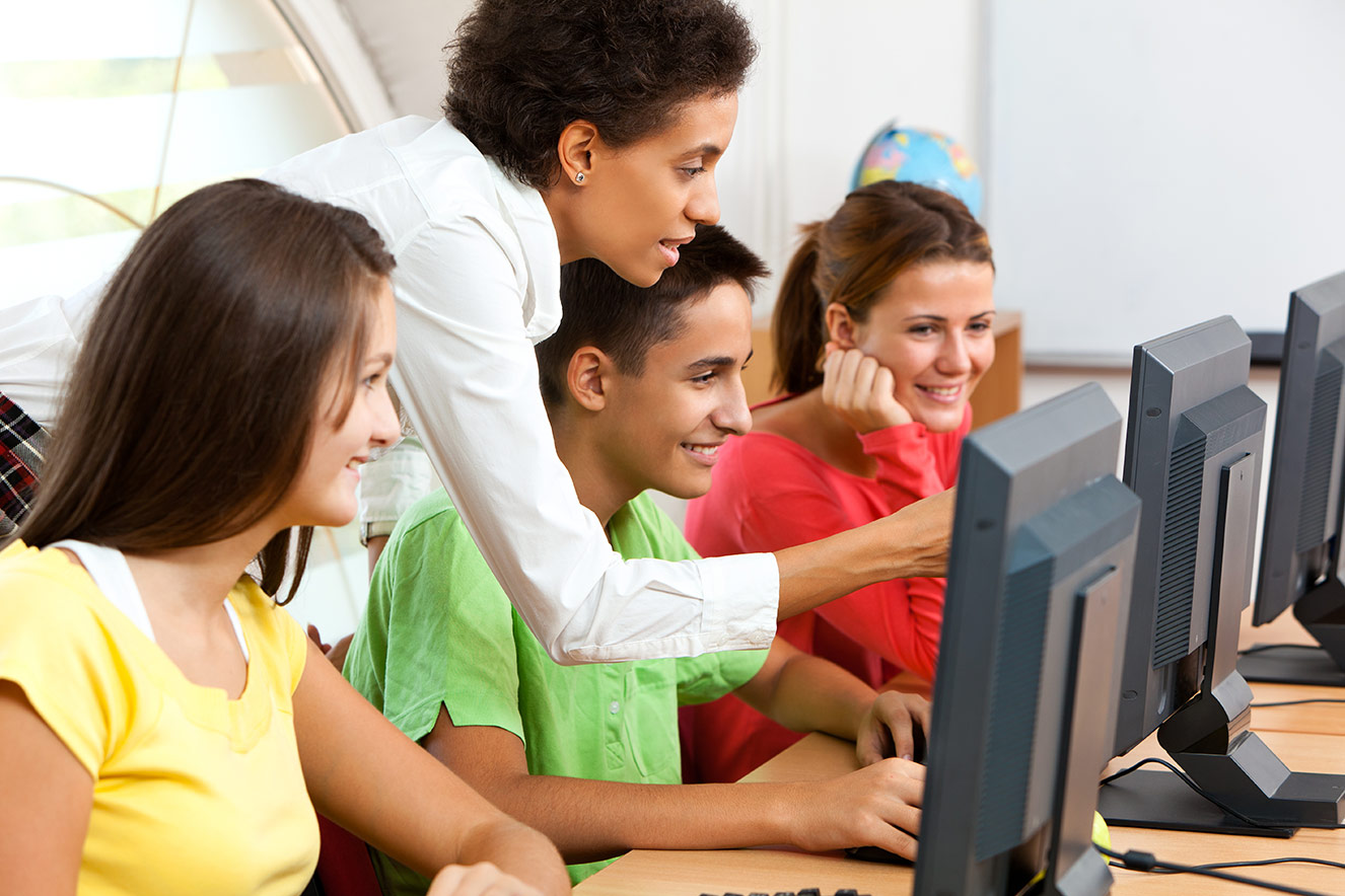 Teacher pointing to a computer screen while 3 students look on in a computer lab