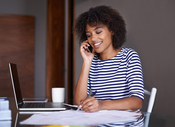 Person wearing a striped shirt, sitting at a table and talking on the phone while taking notes