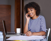 Person wearing a striped shirt, sitting at a table and talking on the phone while taking notes