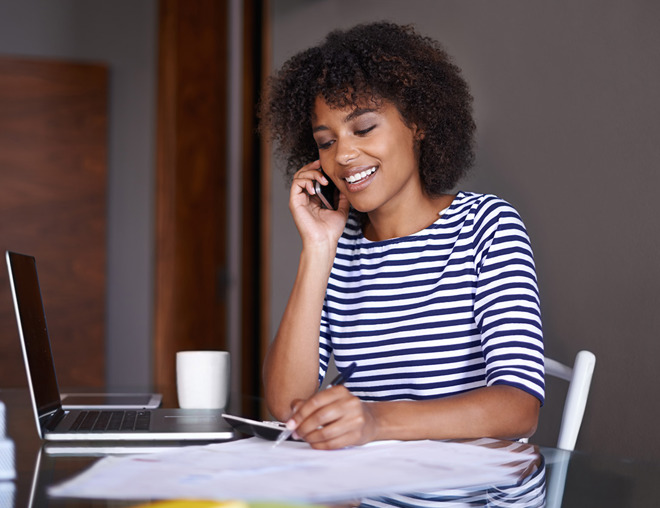 Person wearing a striped shirt, sitting at a table and talking on the phone while taking notes