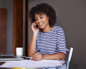 Person wearing a striped shirt, sitting at a table and talking on the phone while taking notes