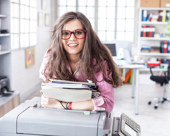 Person wearing glasses, carrying documents, and leaning on a copier in an office
