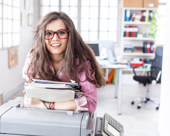 Person wearing glasses, carrying documents, and leaning on a copier in an office