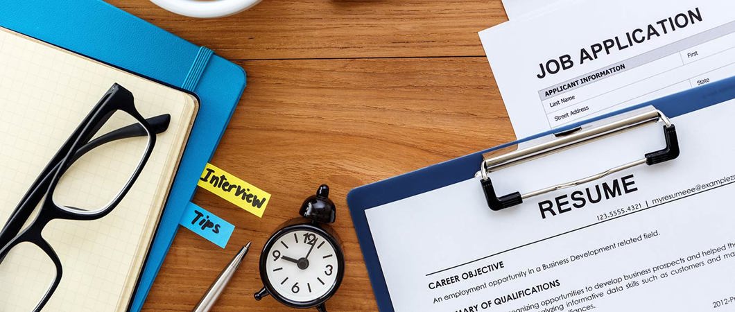 Resume, job application, notebooks, keyboard, coffee and a clock laid out on a desk