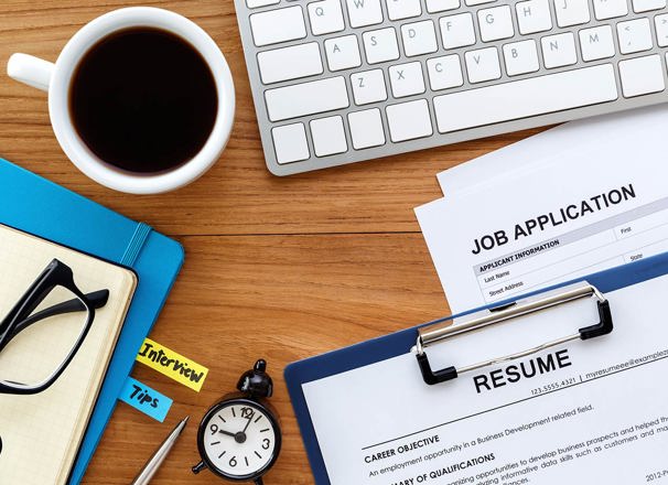 Resume, job application, notebooks, keyboard, coffee and a clock laid out on a desk