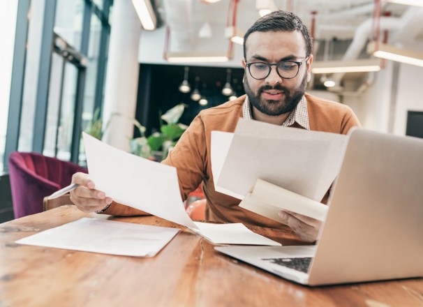 Person sitting at computer and looking through several pieces of paper.