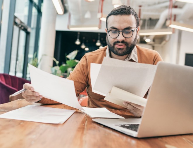 Person sitting at computer and looking through several pieces of paper.