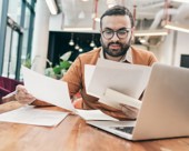 Person sitting at computer and looking through several pieces of paper.