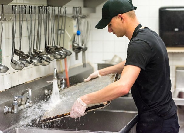 Man washing baking sheet in the sink of a commercial kitchen.