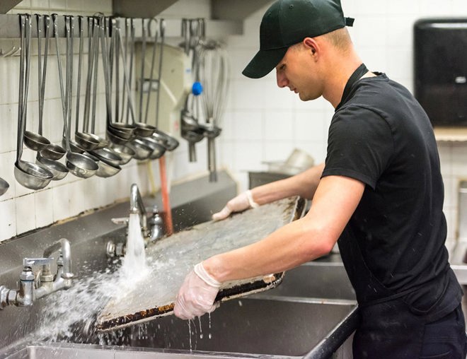 Man washing baking sheet in the sink of a commercial kitchen.