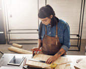 Woman doing woodworking while looking at a tablet in her workshop.