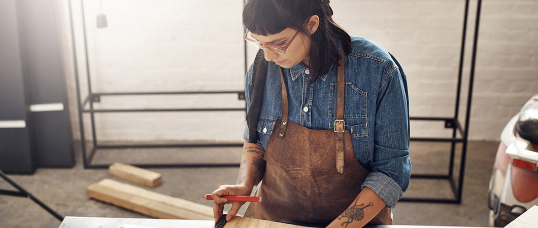 Woman doing woodworking while looking at a tablet in her workshop.