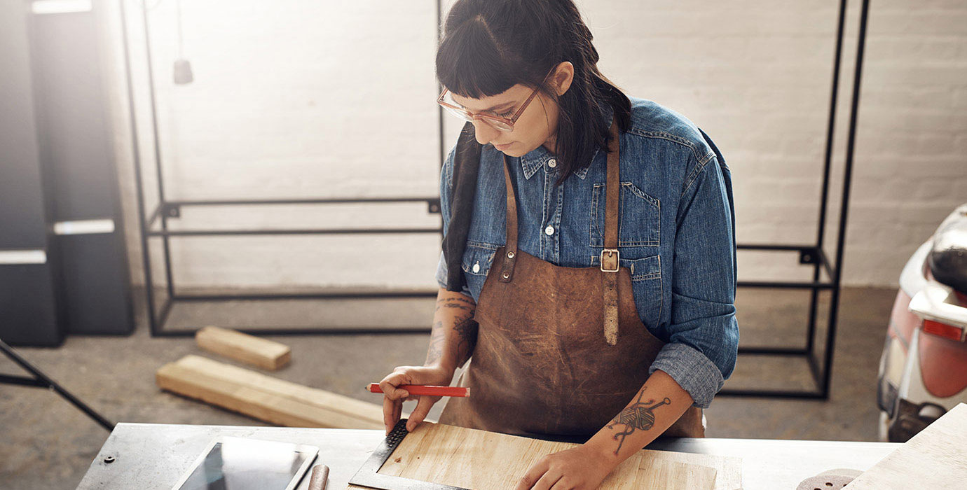 Woman doing woodworking while looking at a tablet in her workshop.