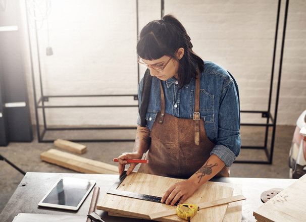Woman doing woodworking while looking at a tablet in her workshop.