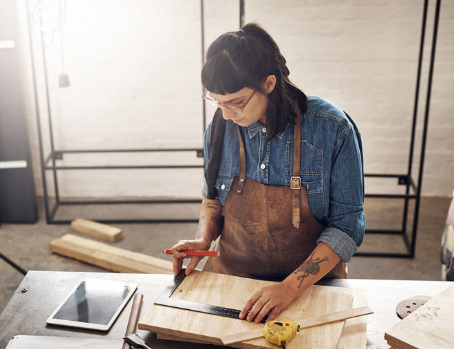 Woman doing woodworking while looking at a tablet in her workshop.