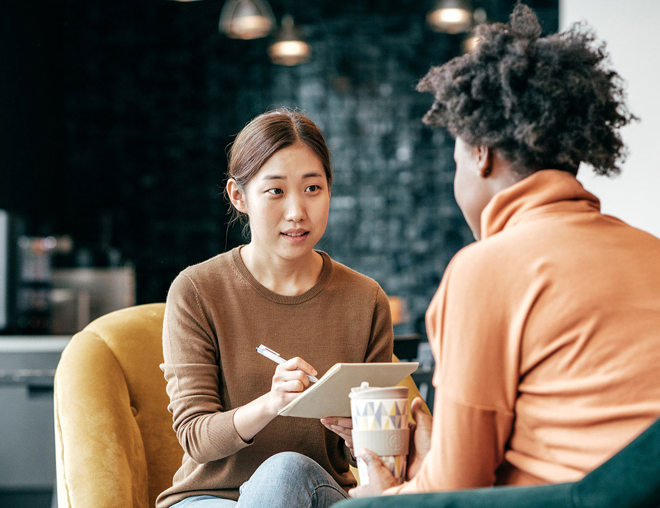 Woman interviewing another woman and taking notes as she speaks.