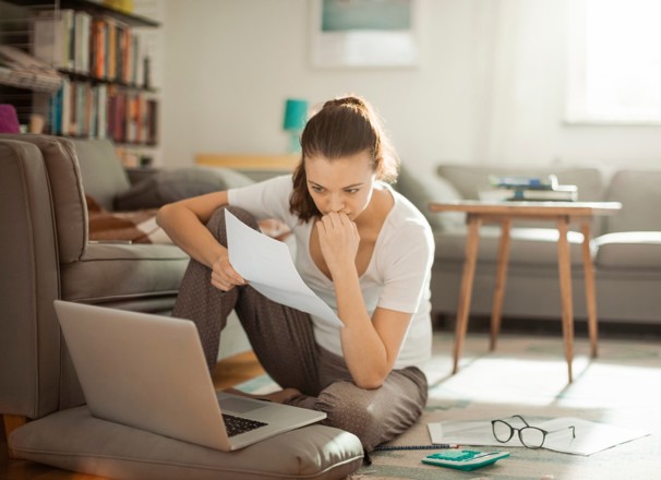 Woman sitting on floor while looking at papers and laptop.