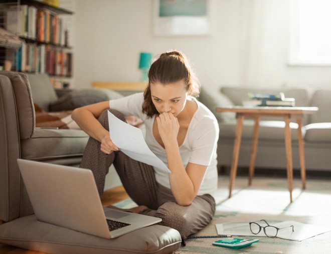 Woman sitting on floor while looking at papers and laptop.
