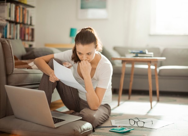 Woman sitting on floor while looking at papers and laptop.