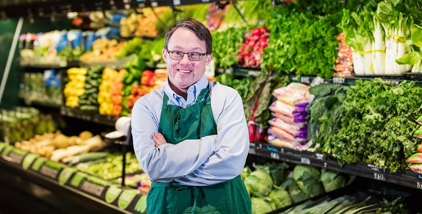 A person smiling in the produce section of a grocery store.