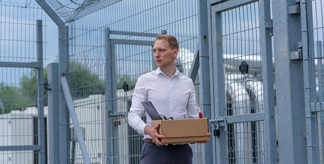 A person carrying a box of personal items standing outside a grey prison fence.