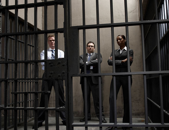 3 people wearing business suits with arms crossed standing inside a prison cell.