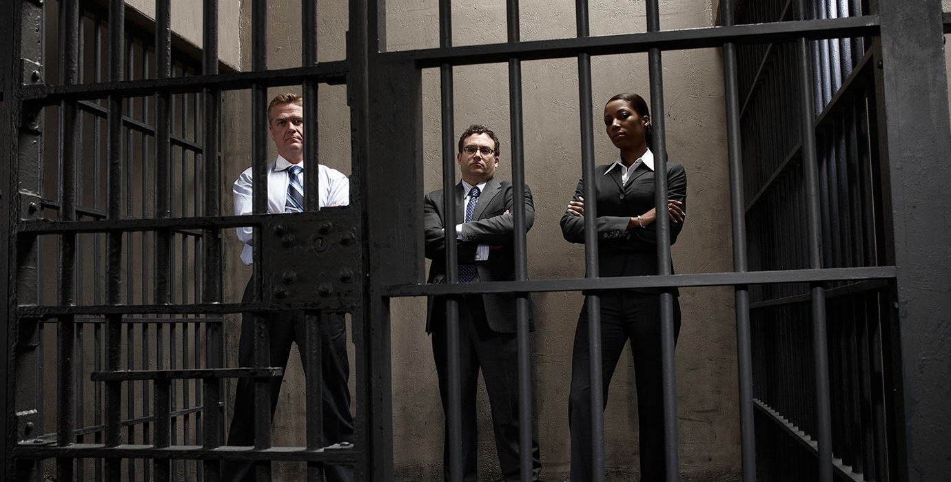 3 people wearing business suits with arms crossed standing inside a prison cell.